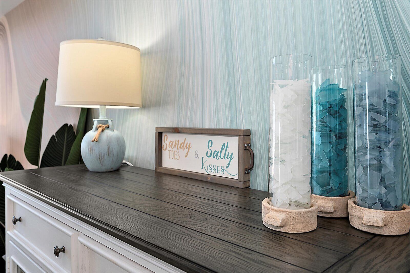 A wooden dresser with a lamp, a framed sign, and three glass containers with crystals.