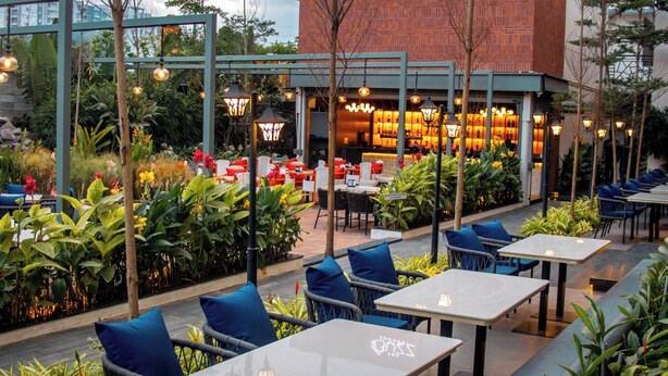 An outdoor seating area with tables and chairs, surrounded by greenery and a building with a sign reading "The Buzz House". - Snap Inn Rooms By The Buzz House (Anekal)