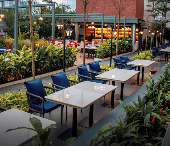 An outdoor seating area with tables and chairs, surrounded by greenery and a building with a sign reading "The Buzz House". - Snap Inn Rooms By The Buzz House (Anekal)
