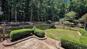 A landscaped garden with a paved patio, a wooden fence, and a variety of shrubs and trees. - Buford Home NEAR everything! (Buford)