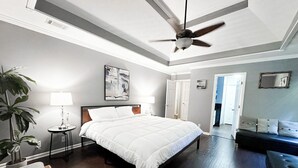 A bedroom with a bed, a ceiling fan, a nightstand, a lamp, a plant, and a view of a hallway. - Buford Home NEAR everything! (Buford)
