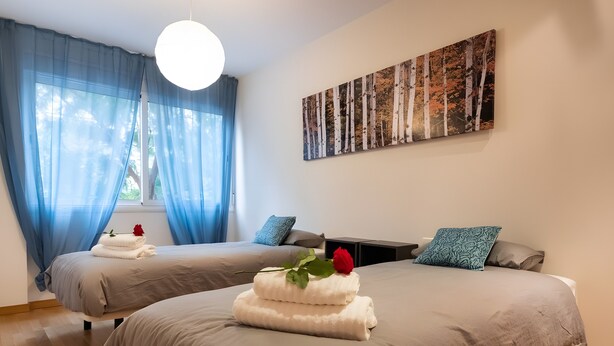 Habitación de hotel con dos camas, un ventanal con cortinas azules, un cuadro colgado en la pared y una lámpara de techo redonda.