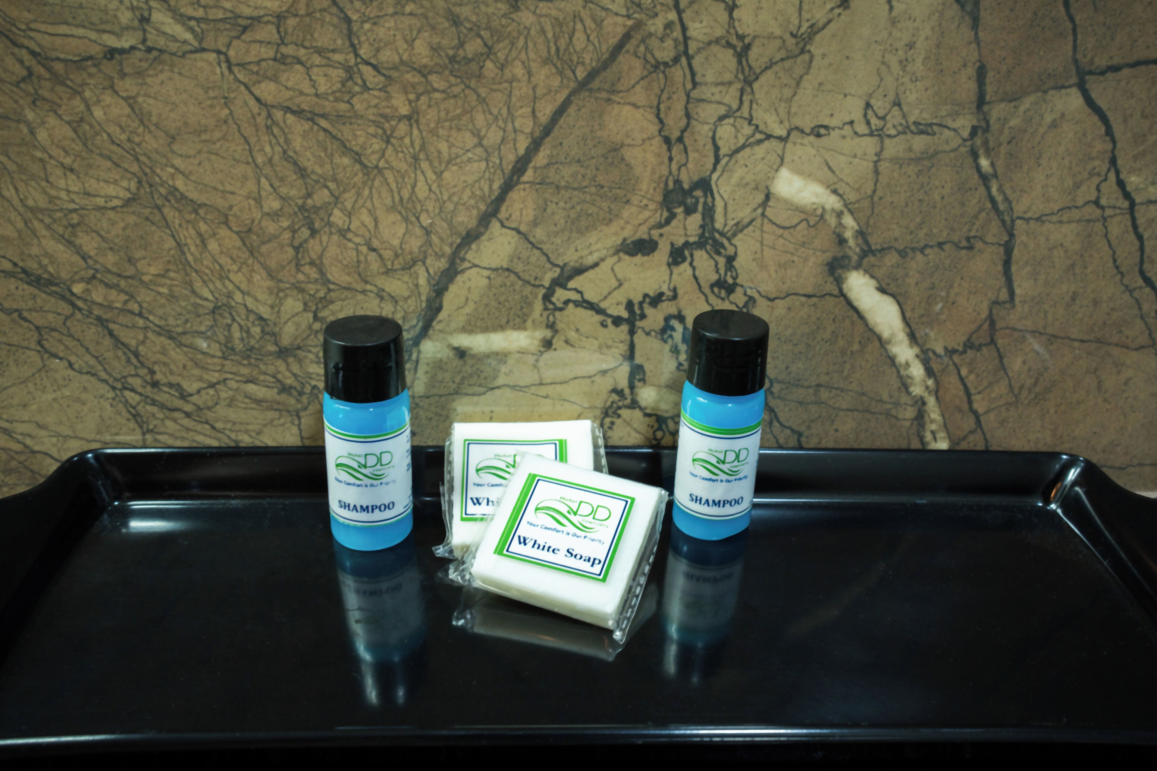 A black tray with three bottles of hotel shampoo and two bars of hotel white soap against a marble wall.