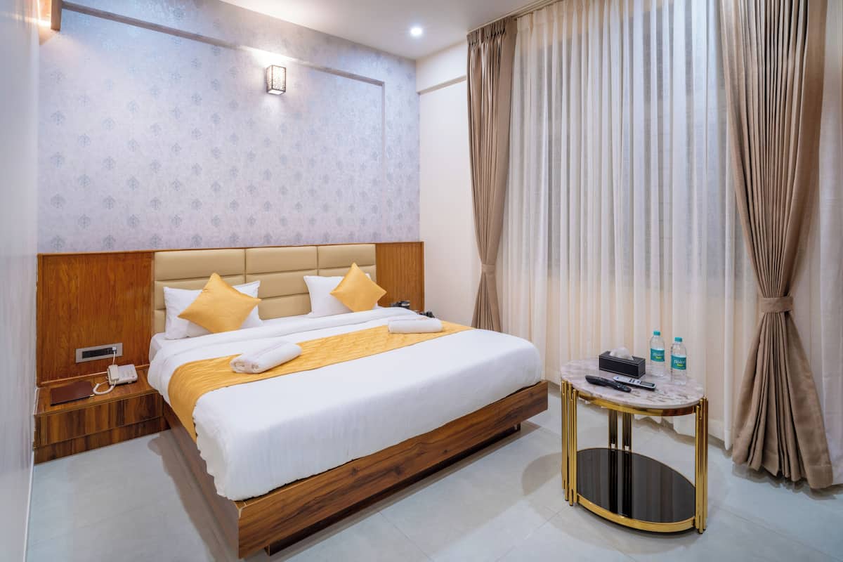 A hotel room with a wooden headboard, a bed with white and yellow bedding, a bedside table with a phone and water bottles, and a round side table with a remote control.