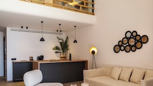 Una sala de estar moderna con un sofá, un sillón blanco, una mesa de centro, una lámpara de pie y una zona de cocina con mostrador e iluminación colgante.