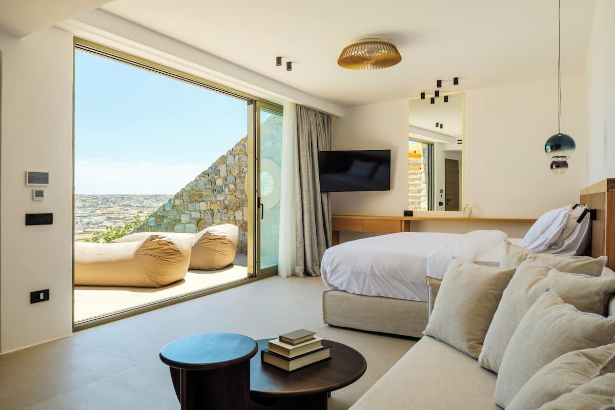 A modern hotel room with a large sliding glass door, a bed, a sofa, a TV, and a small table with books.