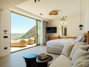 A modern hotel room with a large sliding glass door, a bed, a sofa, a TV, and a small table with books. - Panormos Bay Collection (Mykonos)