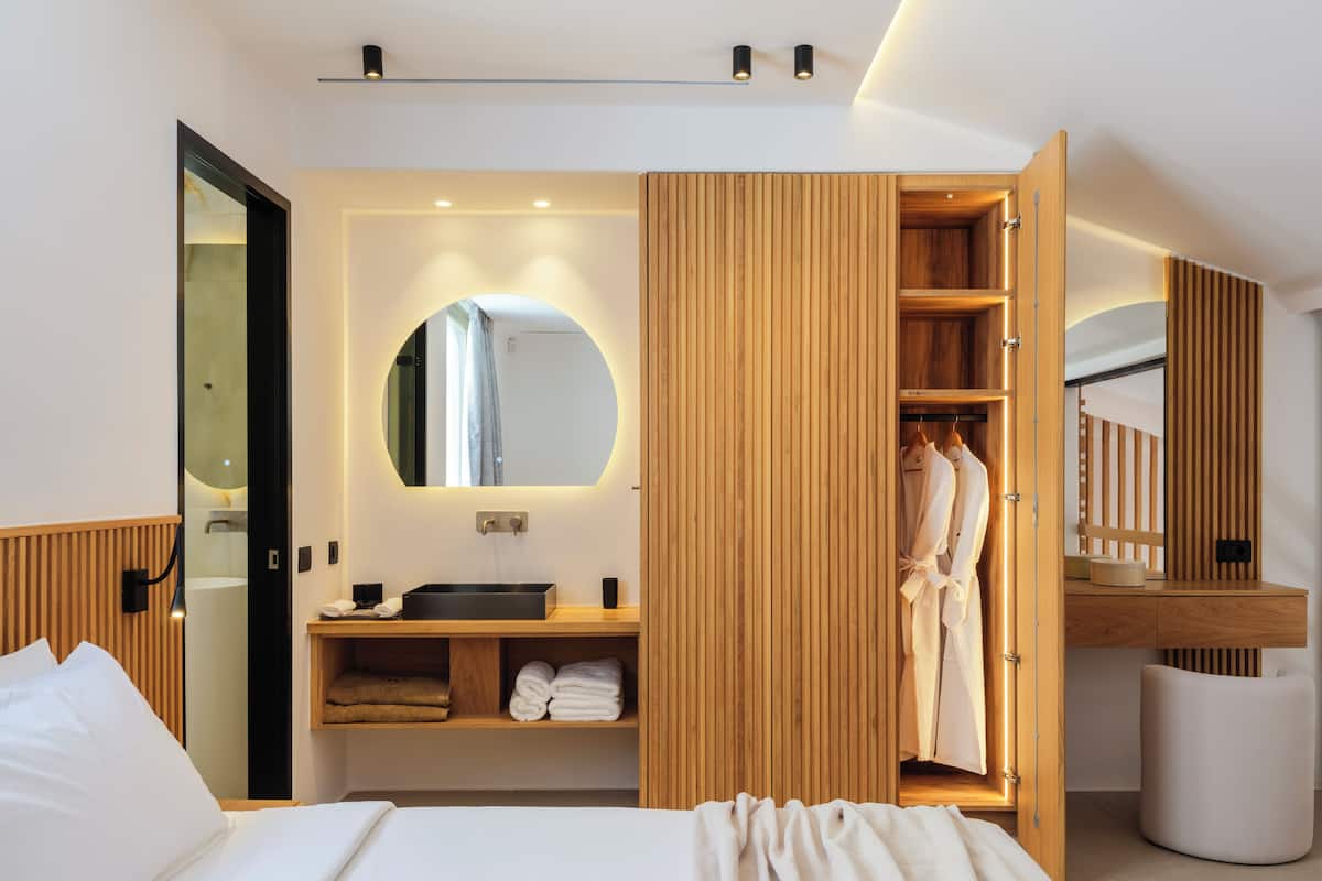 A modern hotel room with a large bed, a wooden wardrobe, a round mirror, a sink, and a robe hanging inside the wardrobe.