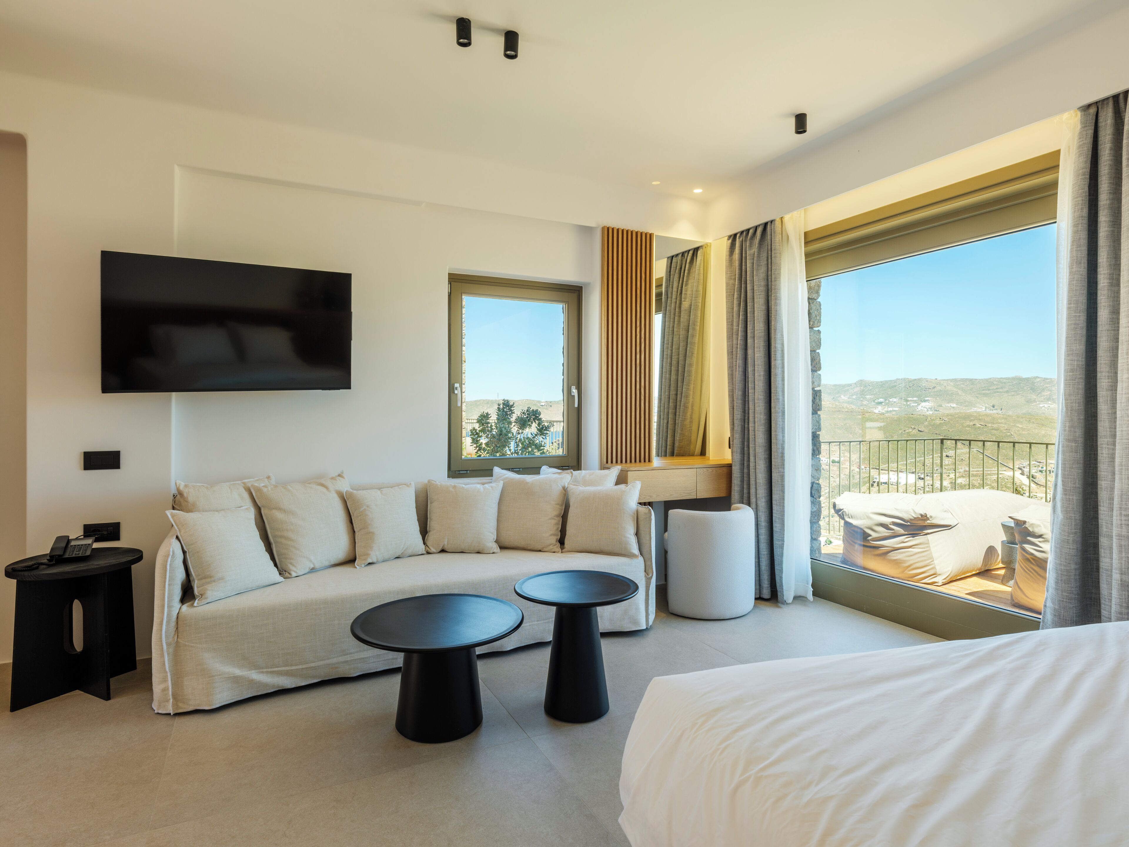 A modern hotel room with a sofa, two round tables, a bed, a TV, and a balcony with a view.
