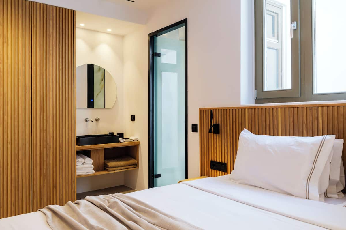 A modern hotel room with a bed, a wooden headboard, a built-in shelf with towels, a round mirror, and a window with natural light.