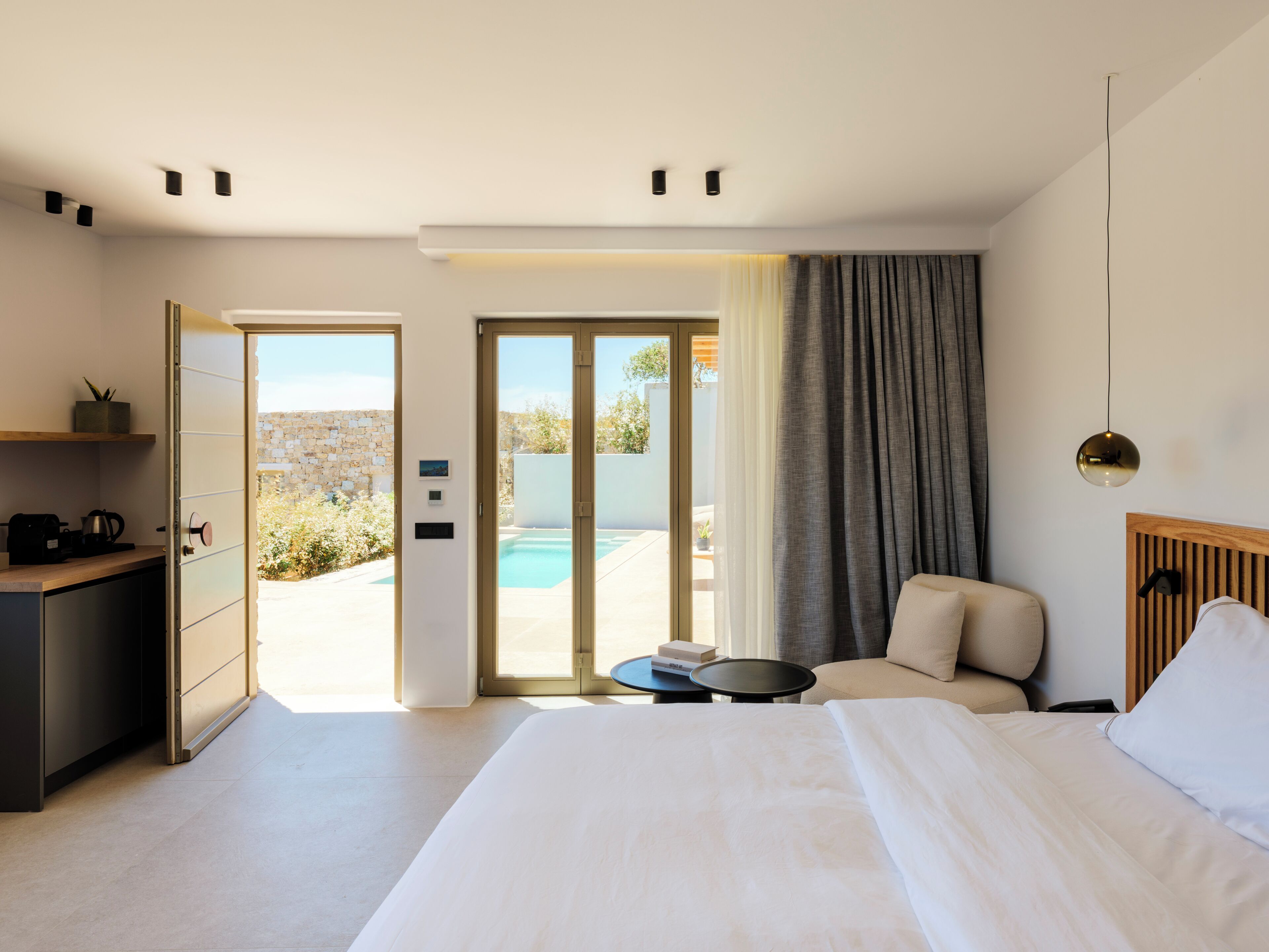 A modern hotel room with a bed, a desk, a chair, a small table, and a view of an outdoor pool and garden.