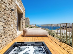 A wooden deck with a hot tub, a stone wall, and a view of the sea. - Panormos Bay Collection (Mykonos)