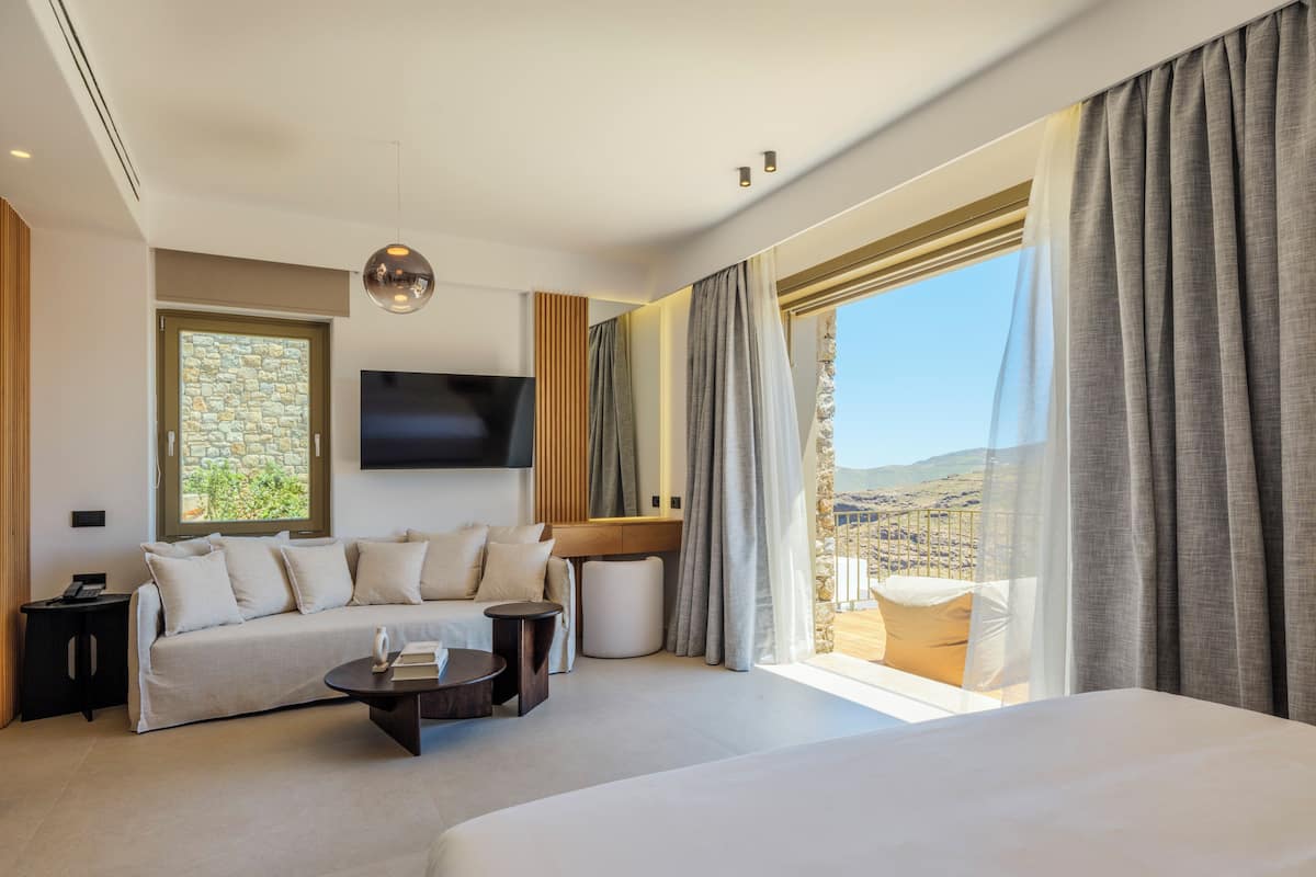 A modern hotel room with a large bed, a sofa, a TV, and a balcony with a view.