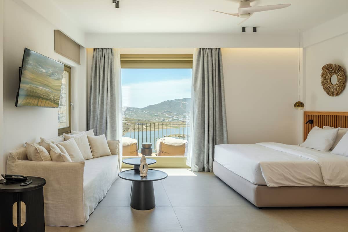 A hotel room with a bed, a sofa, a small table, a ceiling fan, and a view of the outdoors.
