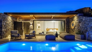 Un área de piscina moderna al aire libre con muros de piedra, una pérgola de madera y un dormitorio con paredes de vidrio que incluye una cama y zona de estar.