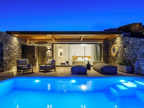 Un espace piscine extérieur moderne, agrémenté de murs en pierre, d’une pergola en bois et d’une chambre à coucher fermée par des baies vitrées, comprenant un lit et un coin salon.