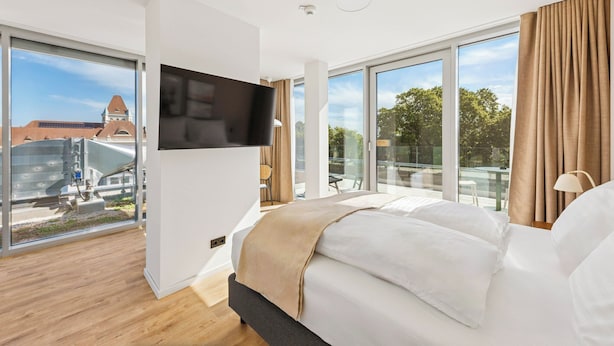 A modern hotel room with a large bed, a flat-screen TV, and a view of a cityscape through large windows.