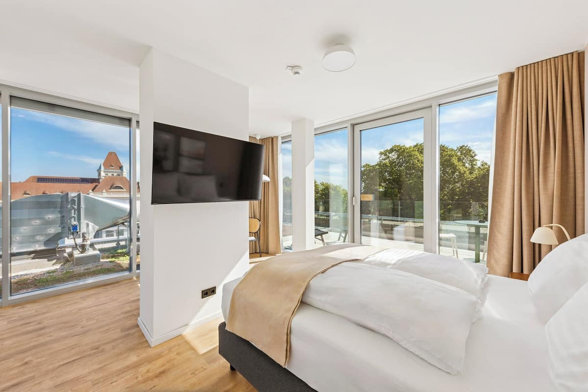 Une chambre d’hôtel moderne dotée d’un grand lit, d’un téléviseur à écran plat et offrant une vue sur le paysage urbain grâce à de grandes fenêtres.