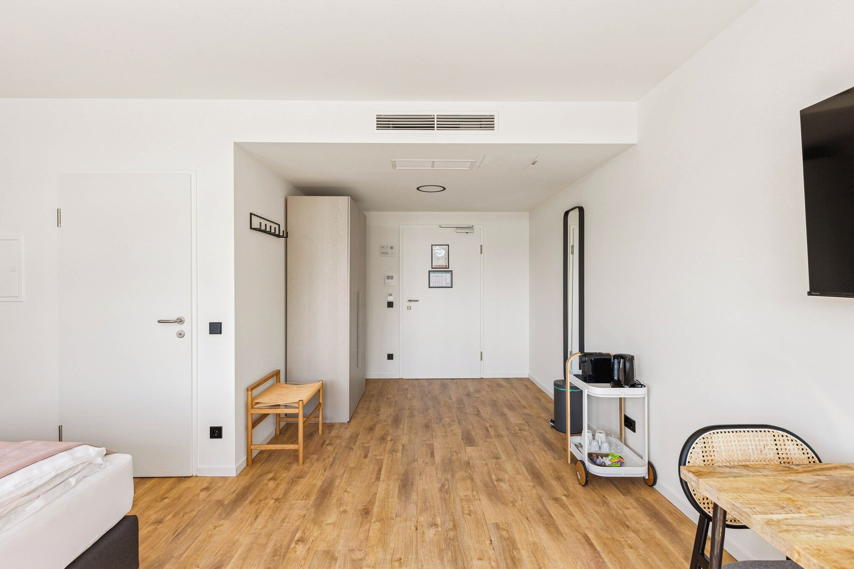 A hotel room with a wooden floor, a bed, a desk, and a chair. There is a door leading to another room and a wall-mounted TV.