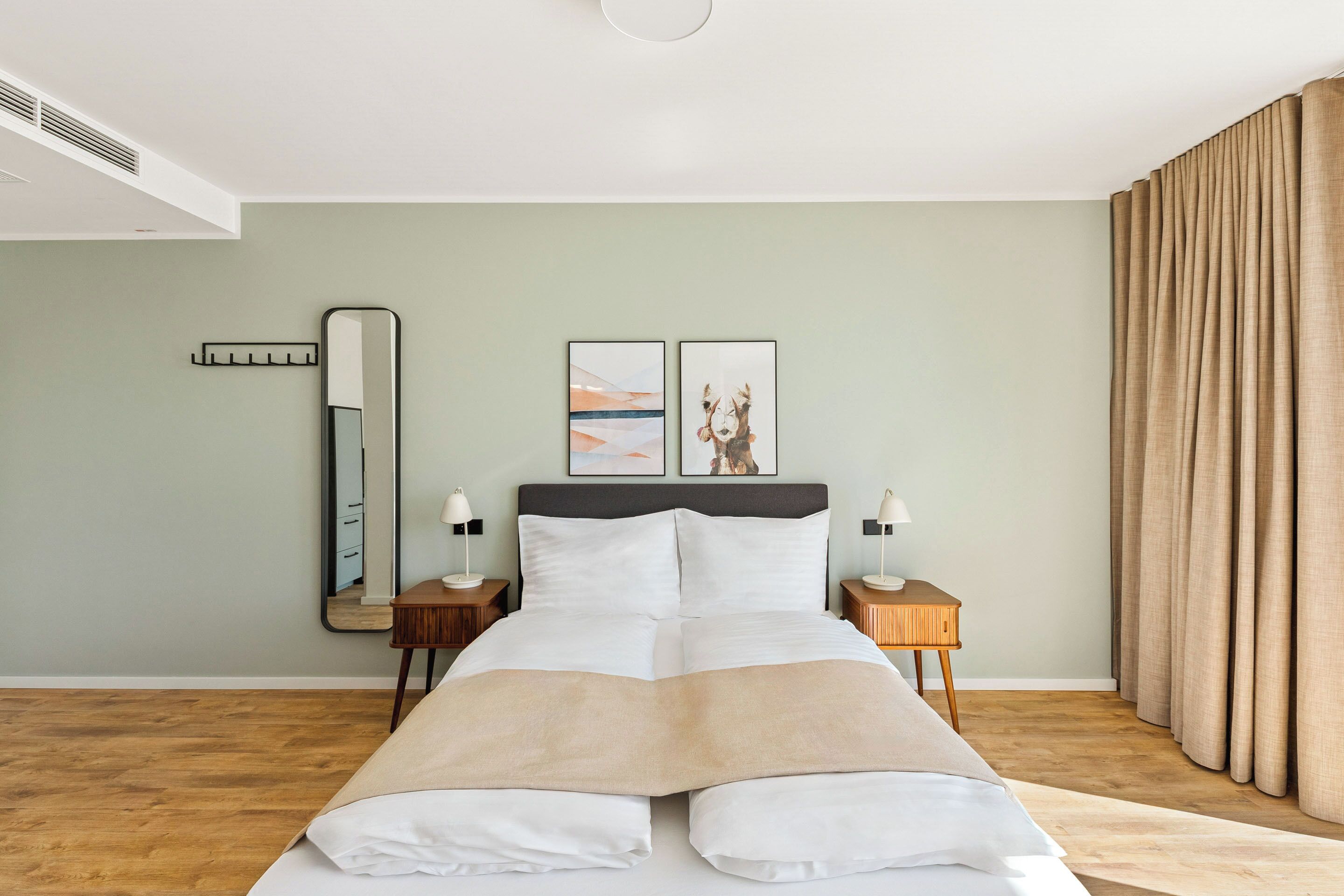 A neatly made bed with a beige throw blanket, a large mirror, two framed artworks on the wall, and a wooden nightstand with a lamp.