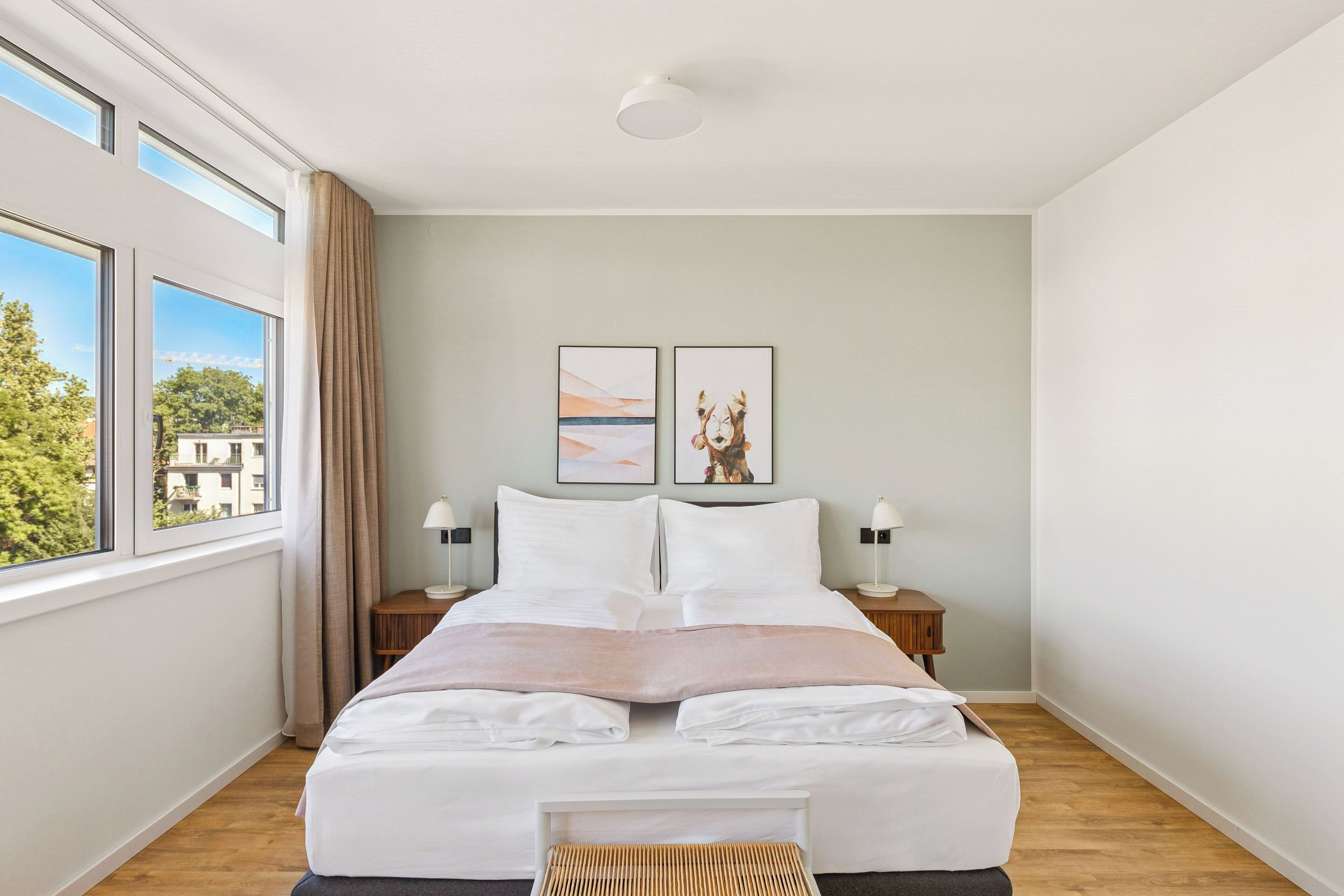 Quarto de hotel moderno com uma cama grande, duas mesas de cabeceira com luminárias, uma janela com cortinas e duas imagens emolduradas na parede.