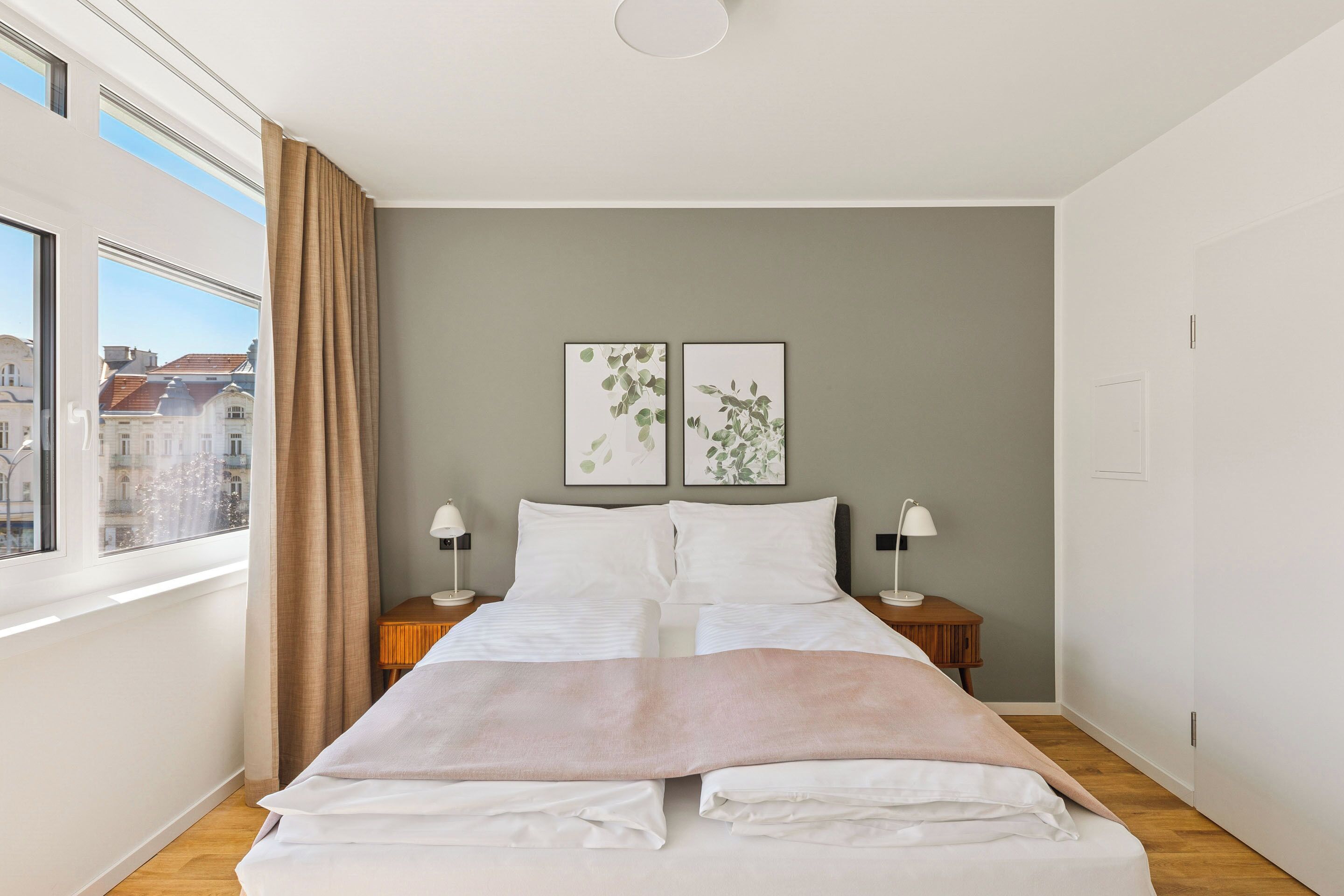 Quarto moderno com uma cama grande, duas mesas de cabeceira com luminárias, uma janela com cortinas e duas molduras com quadros na parede.