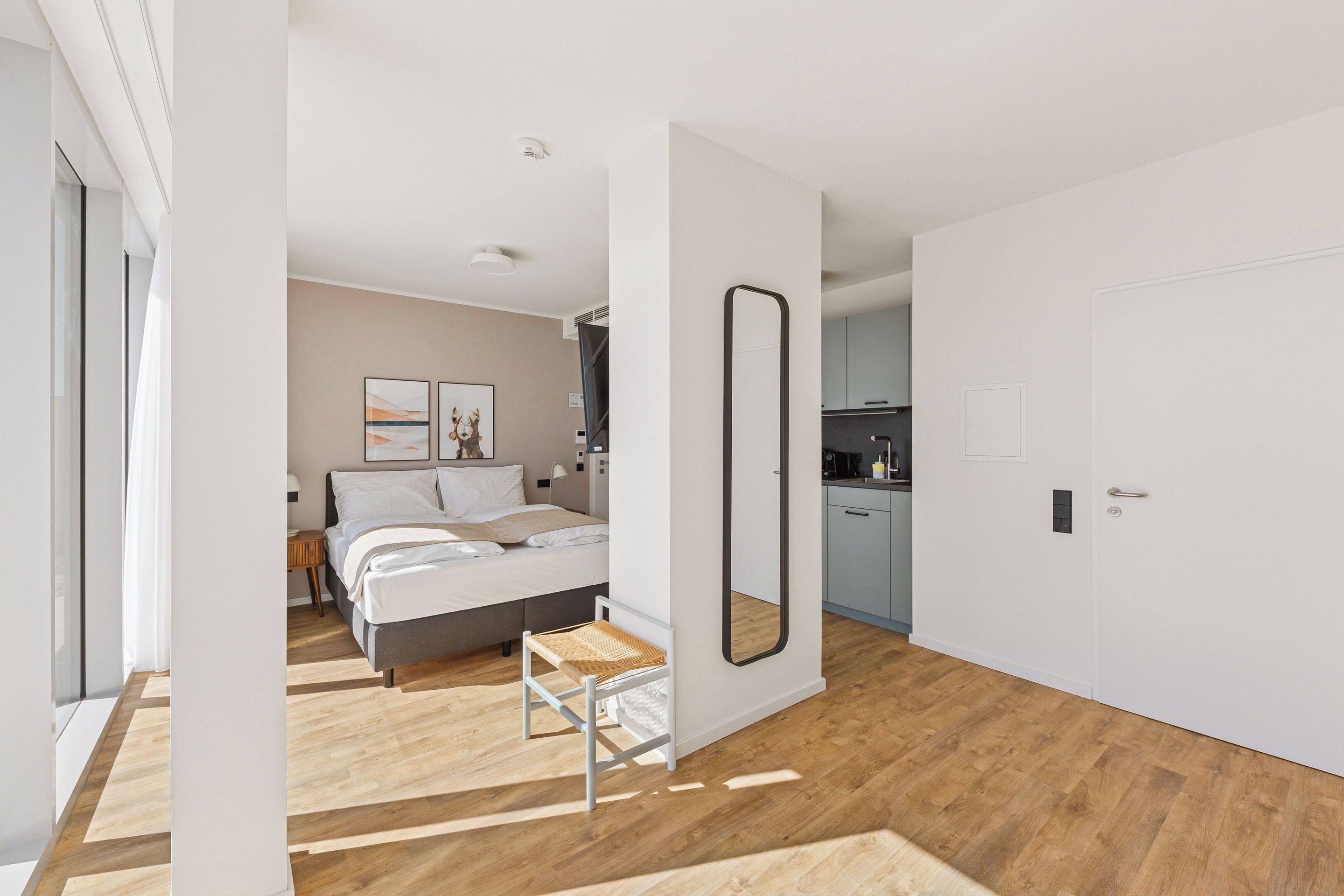 Een moderne slaapkamer met een bed, nachtkastje en een kleine stoel. Er hangt een grote spiegel aan de muur en een deur naar de badkamer.