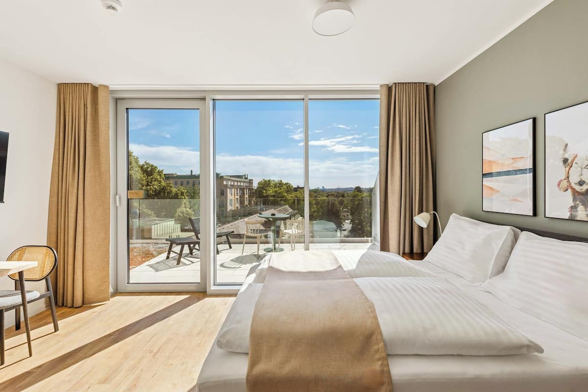 Une chambre d’hôtel moderne dotée d’un grand lit, d’un balcon avec une table et des chaises, et offrant une vue sur le paysage urbain.