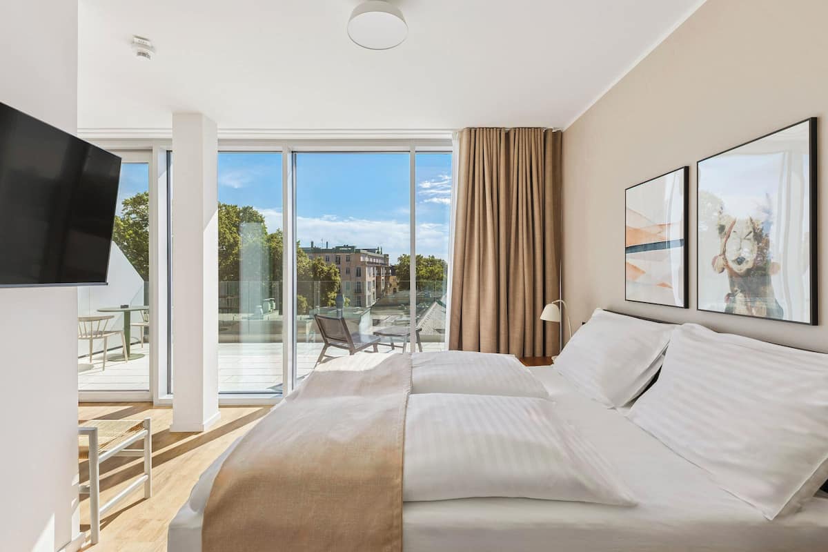 Une chambre d’hôtel moderne dotée d’un grand lit, d’une télévision et d’un balcon offrant une vue sur les bâtiments et les arbres.