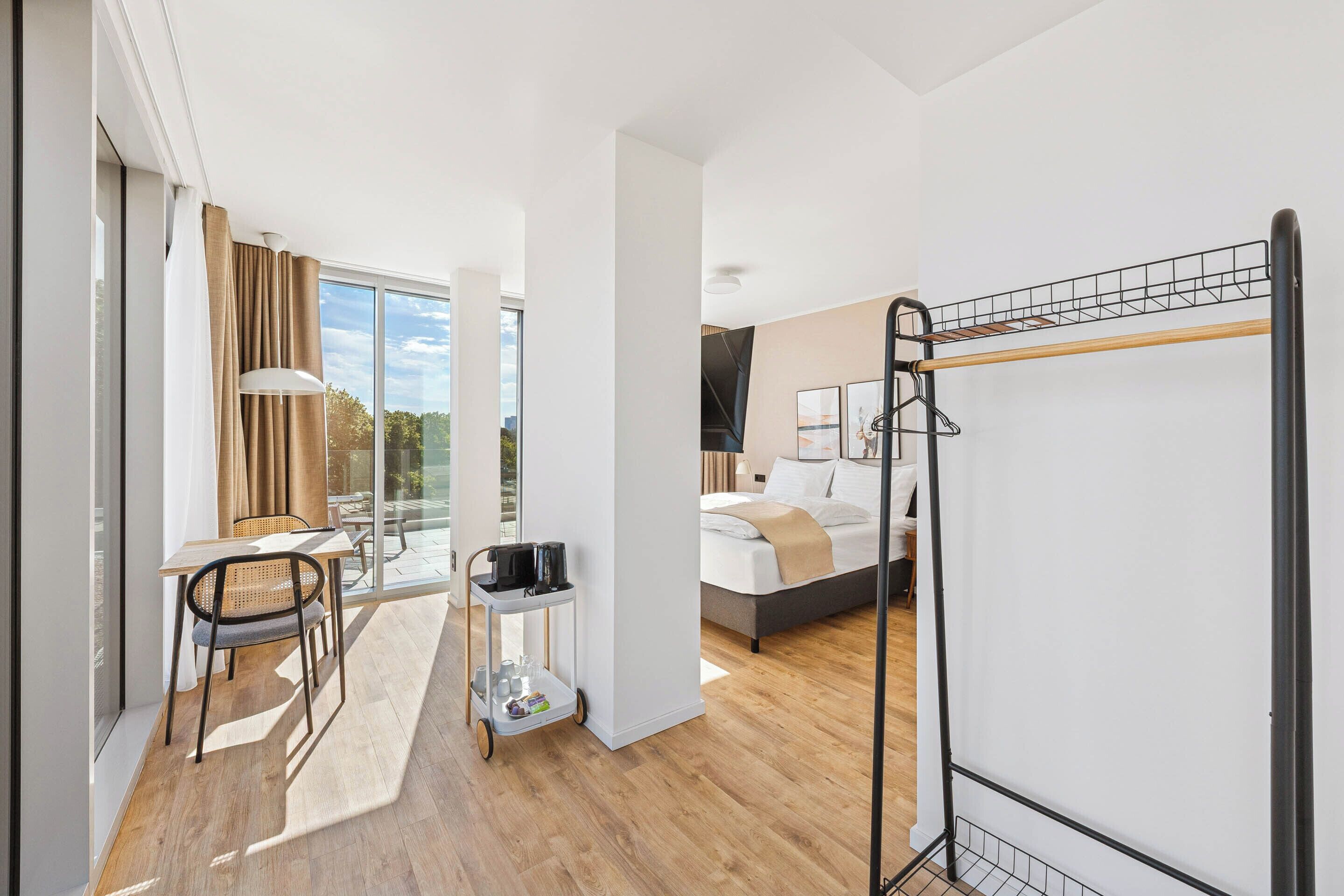 A modern hotel room with a bed, a desk with a chair, a small table with a tray, and a balcony with a view.