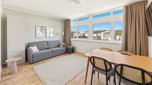 A modern living room with a grey sofa, a dining table with chairs, a small round table, and a view of the city through large windows.