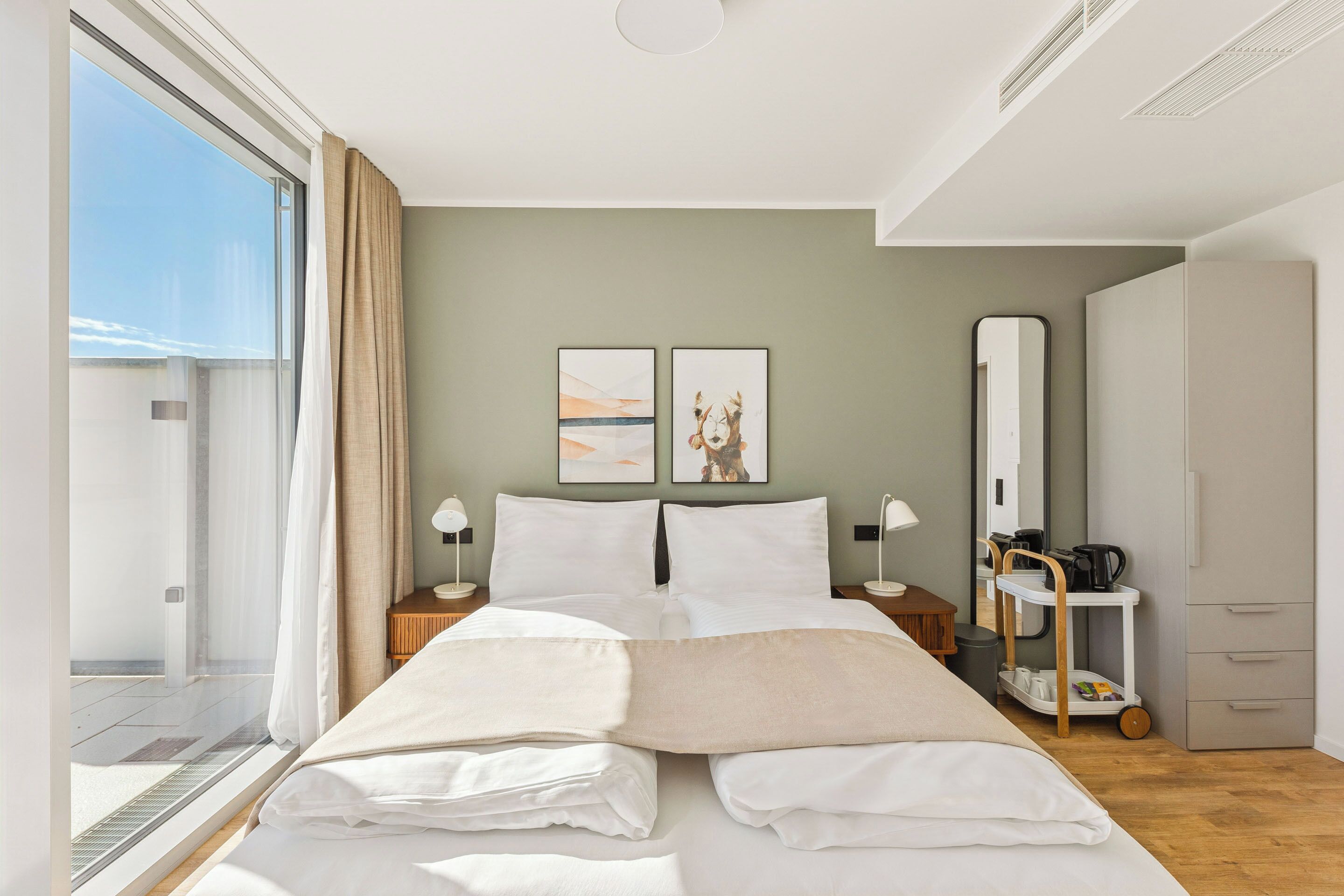 A modern hotel room with a large bed, a bedside table with a lamp, a wardrobe, and a view of the outdoors.
