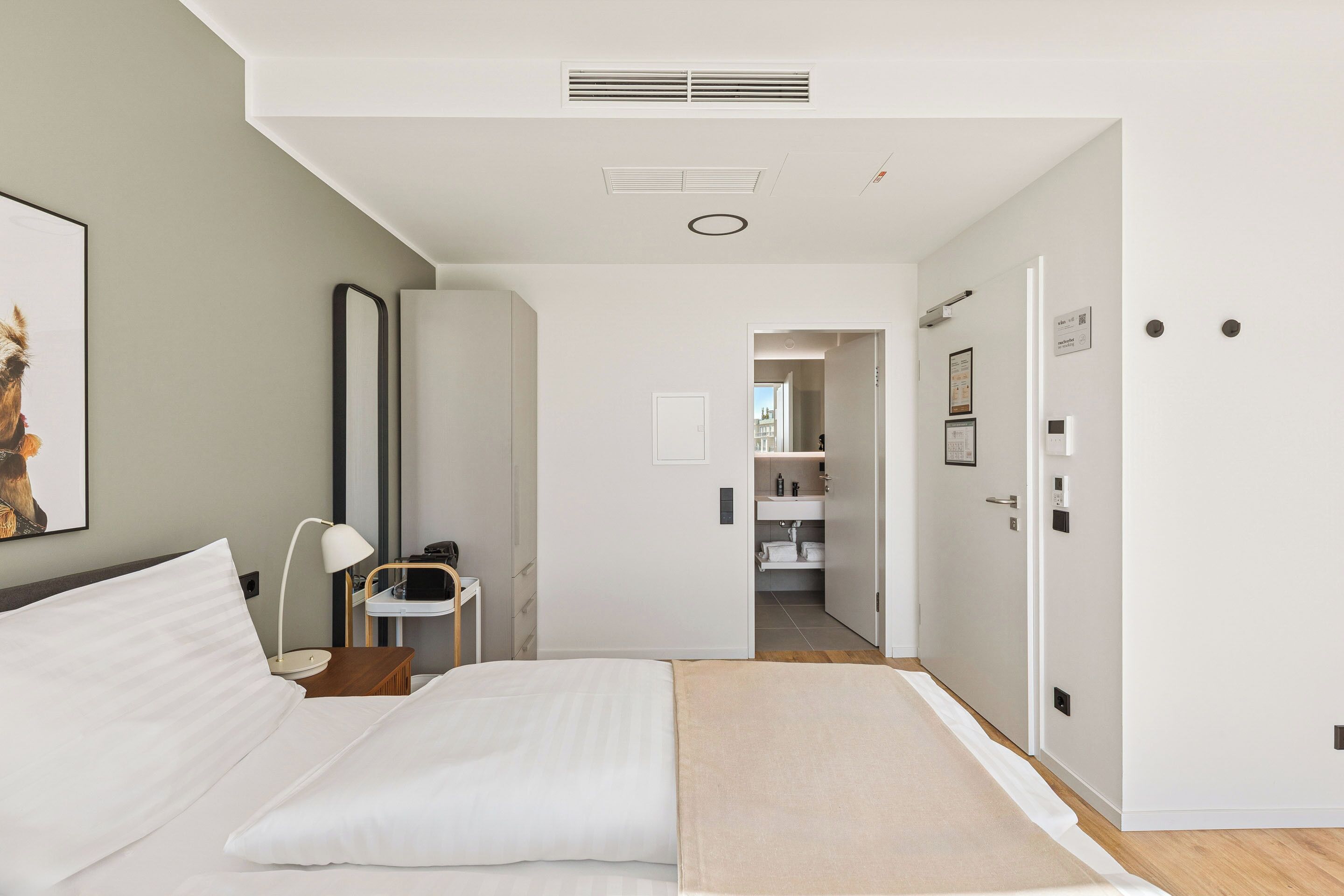 A modern hotel room with a bed, bedside table, lamp, and a view into the bathroom and hallway.