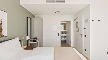 A modern hotel room with a bed, bedside table, lamp, and a view into the bathroom and hallway.