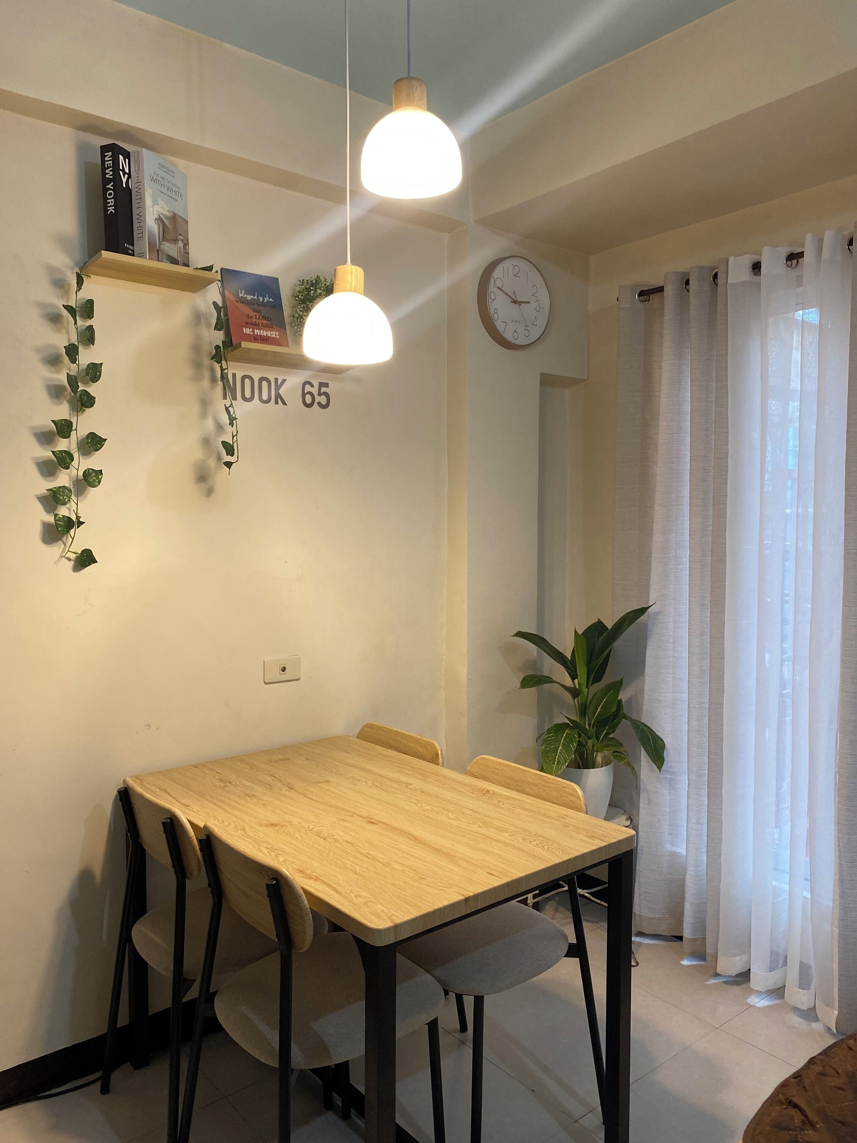 Uma sala de jantar com uma mesa e cadeiras de madeira, um lustre, um relógio e um vaso com planta.