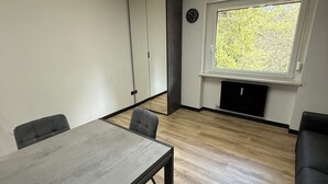 Una habitación moderna con piso de madera, una mesa gris y dos sillas grises. Hay una ventana con vista a árboles y un reloj en la pared.