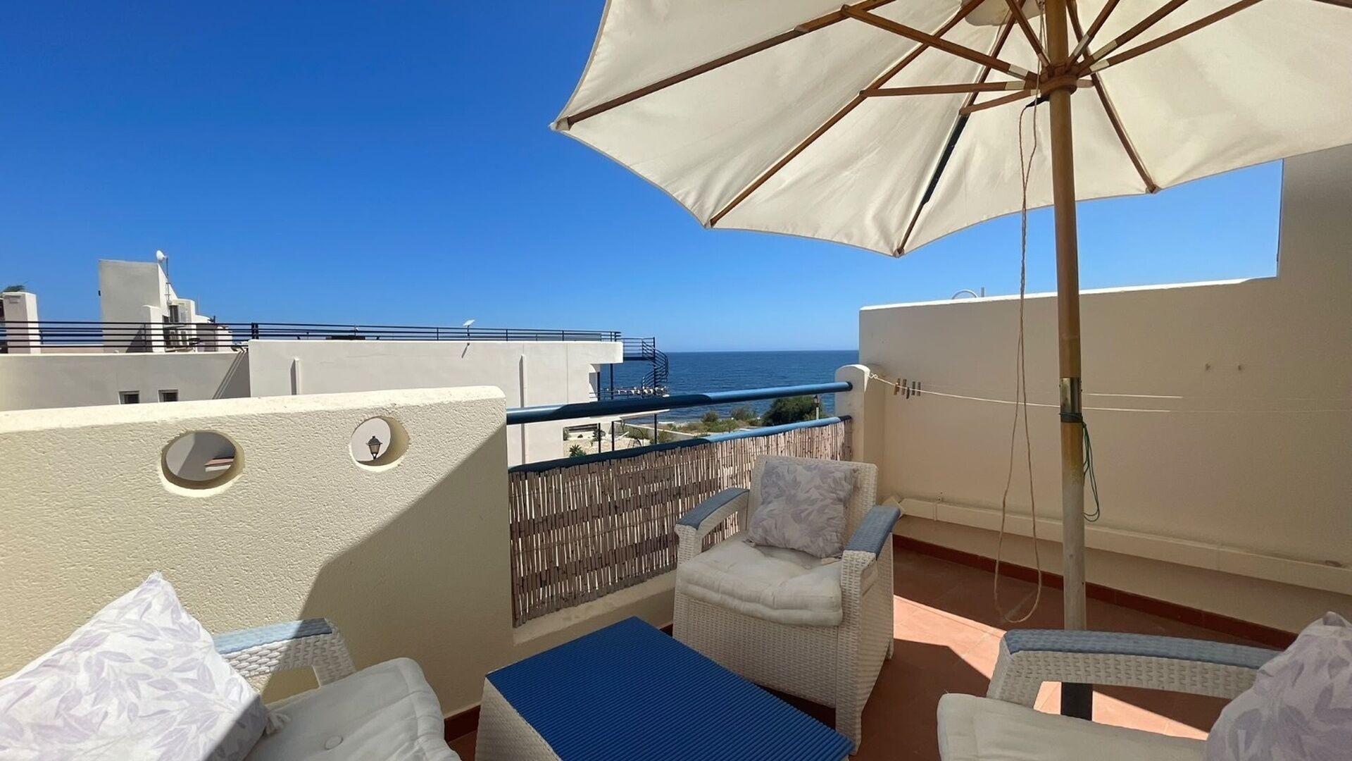 Un balcón con sombrilla blanca, mobiliario de mimbre y vistas al mar.