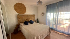 A bedroom with a wooden headboard, a bed with a textured blanket, two pillows, a round woven wall hanging, and a view through a window with curtains. - Casa Luna ;) ... Semi-detached house just a few steps from the ocean 🌊 (Cuevas del Almanzora)