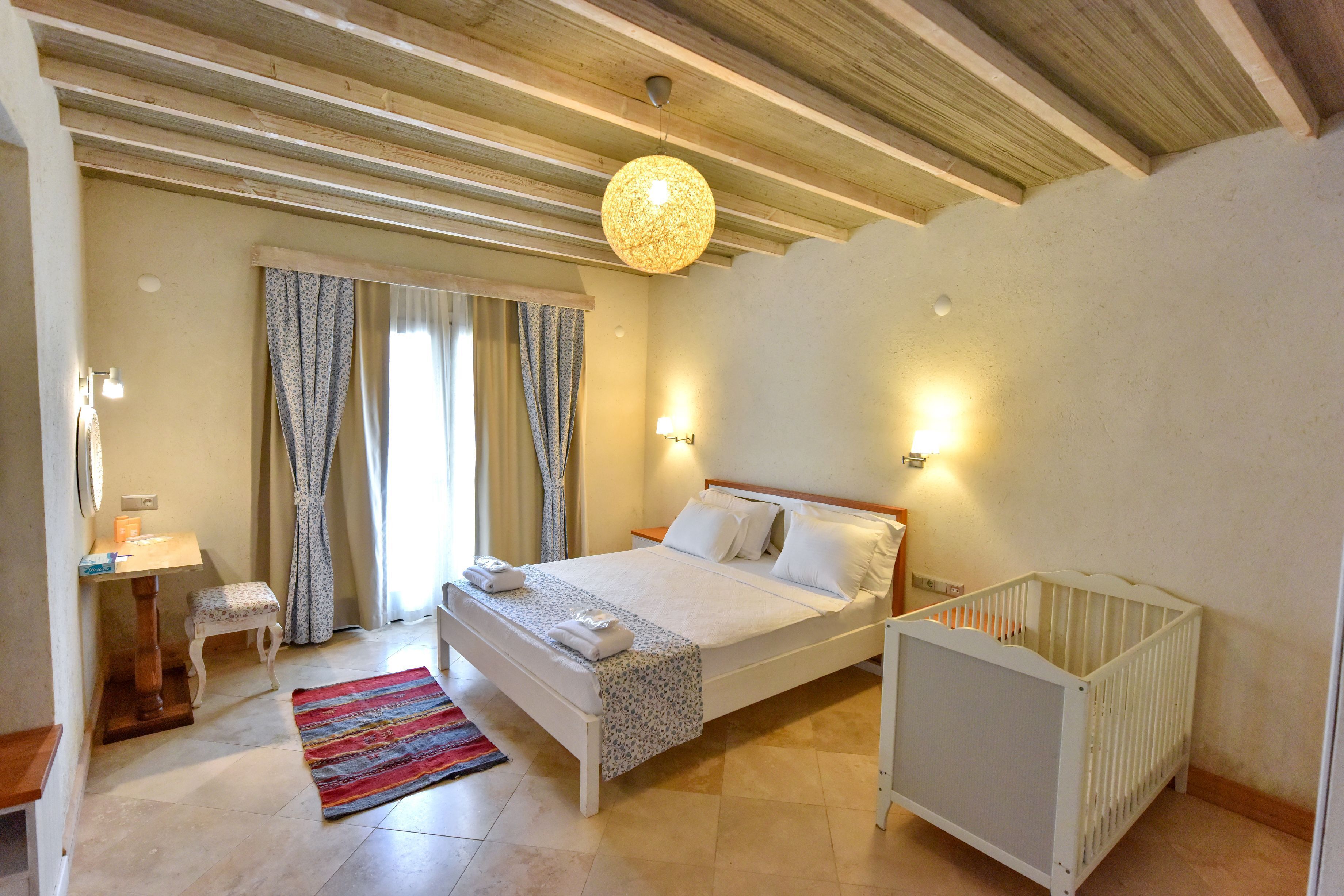 A bedroom with a bed, a crib, a bedside table, a lamp, and a window with curtains.