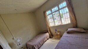 A room with two single beds, a standing fan, a small table with a lamp, and a window with metal bars. - Family-friendly home from home located close to beach, bars and restaurants (Bridgetown)
