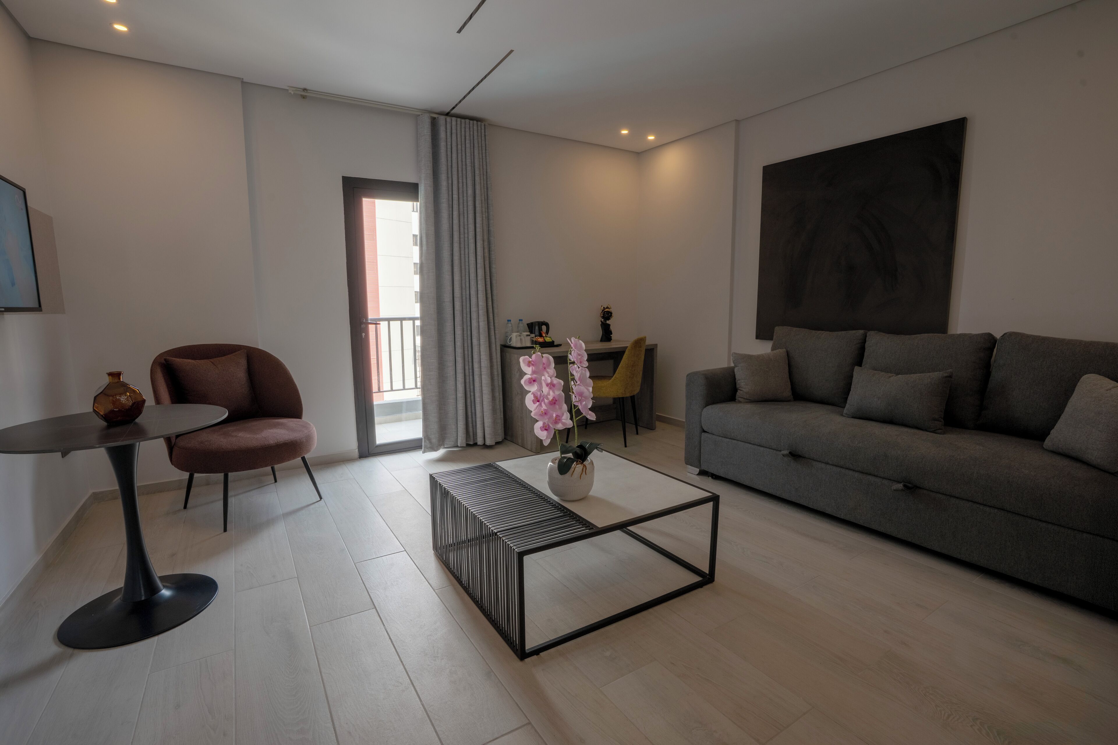 A modern living room with a grey sofa, a round table with a vase of pink flowers, a brown armchair, and a glass coffee table.