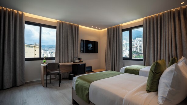 A hotel room with a bed, a desk, a chair, a TV, and a view of the city. - Beverly Beach Hotel (Jounieh)