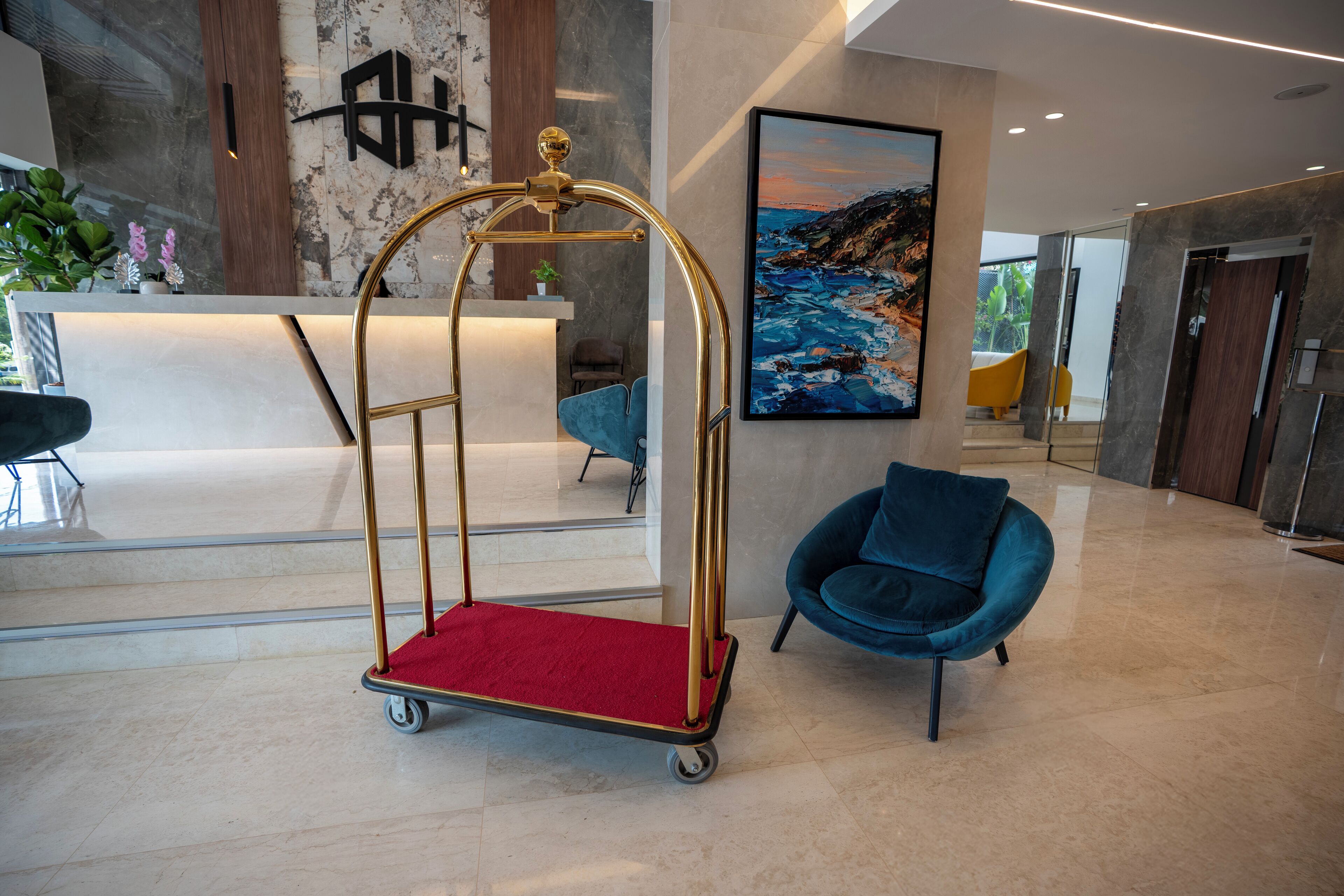 Un hall d’entrée avec un comptoir de réception, un chariot à roulettes à cadre doré surmonté d’un tapis rouge, une chaise bleue et un grand tableau représentant un paysage côtier.