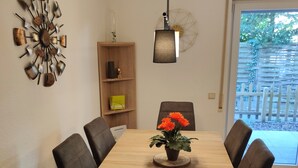 Un comedor con una mesa de madera, seis sillas, una planta en maceta con flores naranjas, un reloj de pared, un estante con objetos decorativos y una lámpara colgante.