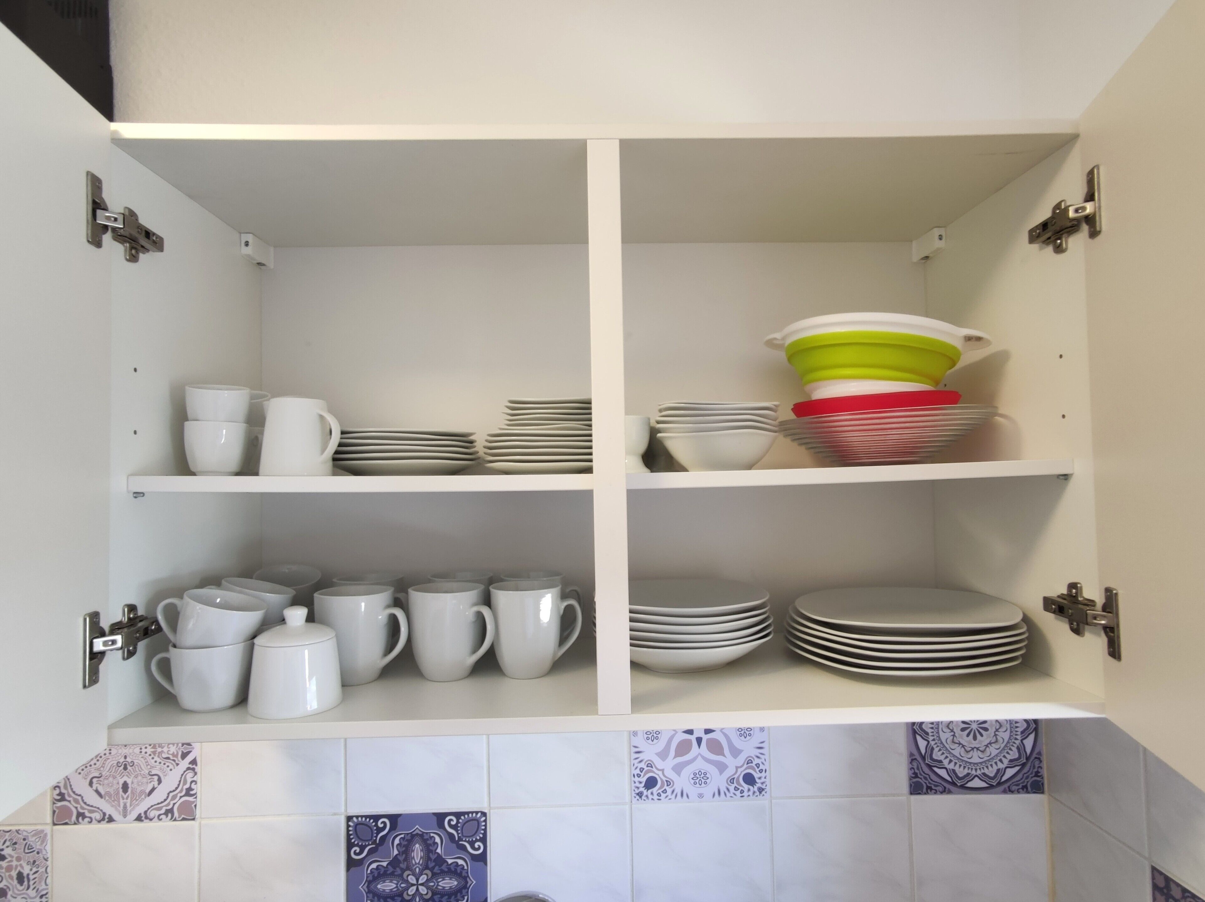 A kitchen cabinet with white shelves, holding white and colored dishes, and a tiled backsplash with patterned tiles.
