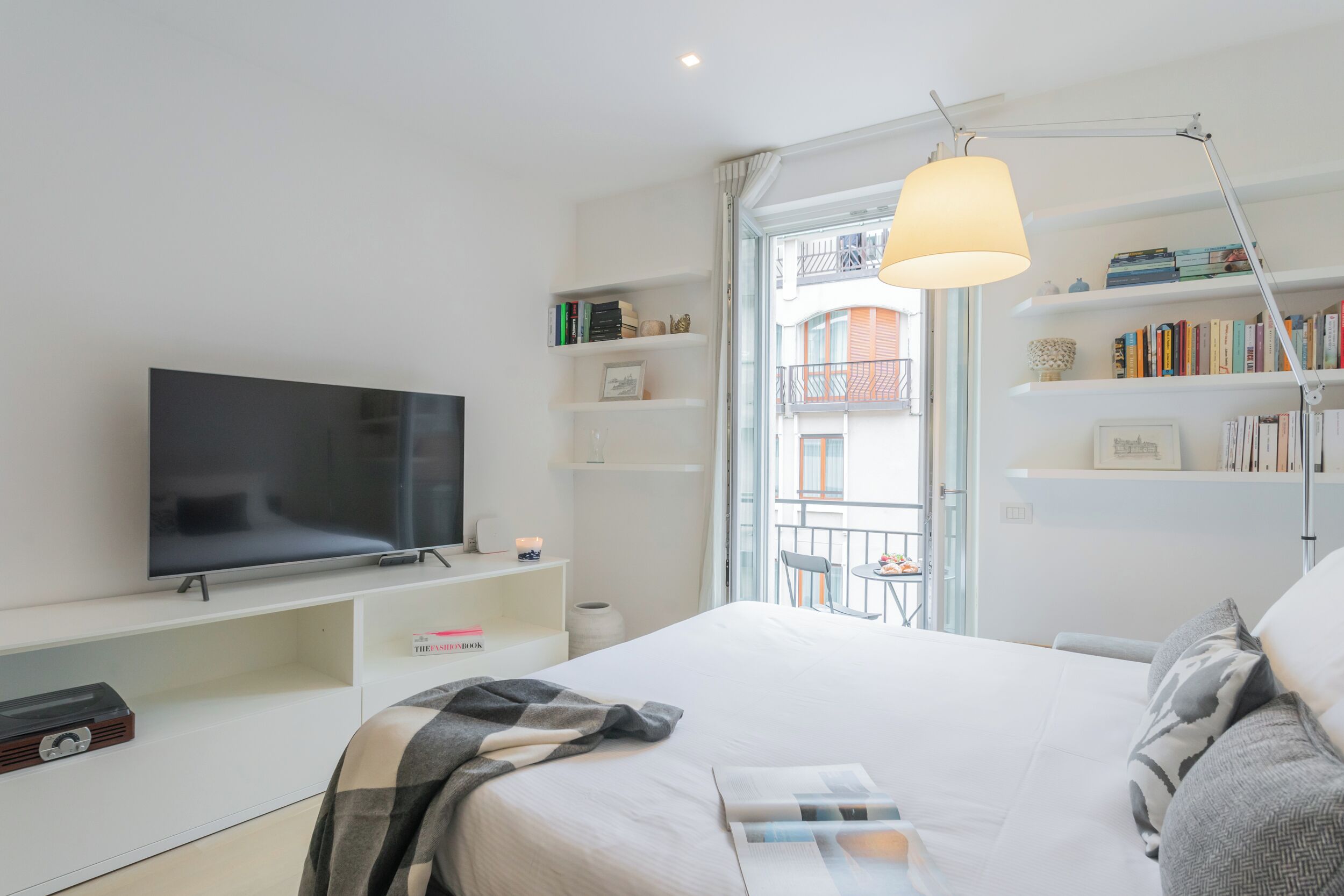 A modern hotel room with a large flat-screen TV, a comfortable bed, a bedside table, a bookshelf, and a balcony with a view of the city.