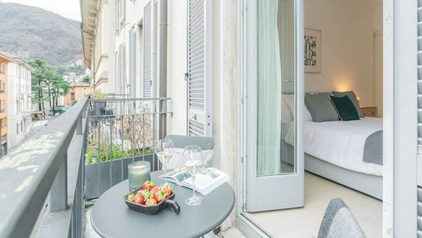 A balcony with a table set for two, a bed, and a view of the city and hills. - Central & White Apartment by Rent all Como (Como)