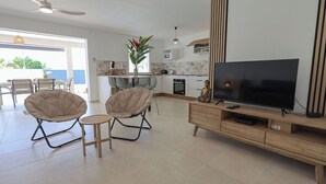 A modern living room with a flat-screen TV, two cushioned chairs, a small round table, and a kitchen area with a dining set.