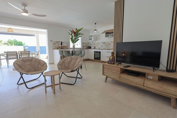 Una sala moderna con un televisor de pantalla plana, dos sillones cómodos, una mesita redonda y una cocina con juego de comedor.