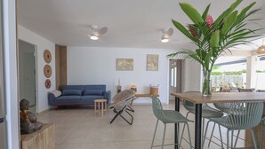 A modern living room with a blue sofa, a dining table with chairs, ceiling fans, and a large plant.